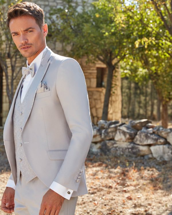 Bräutigam in einem eleganten, hellen Hochzeitsanzug mit Fliege und Weste, stehend im Freien vor einer rustikalen Steinmauer im Sonnenschein.