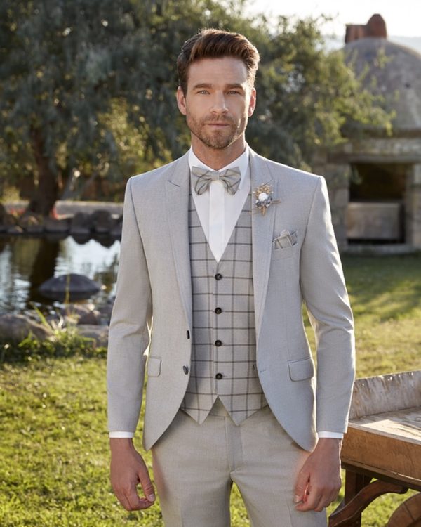 Bräutigam in einem hellen, sommerlichen Dreiteiler mit kariertem Westenmuster, Fliege und Sakko, posierend im Freien vor einer Wasserlandschaft.