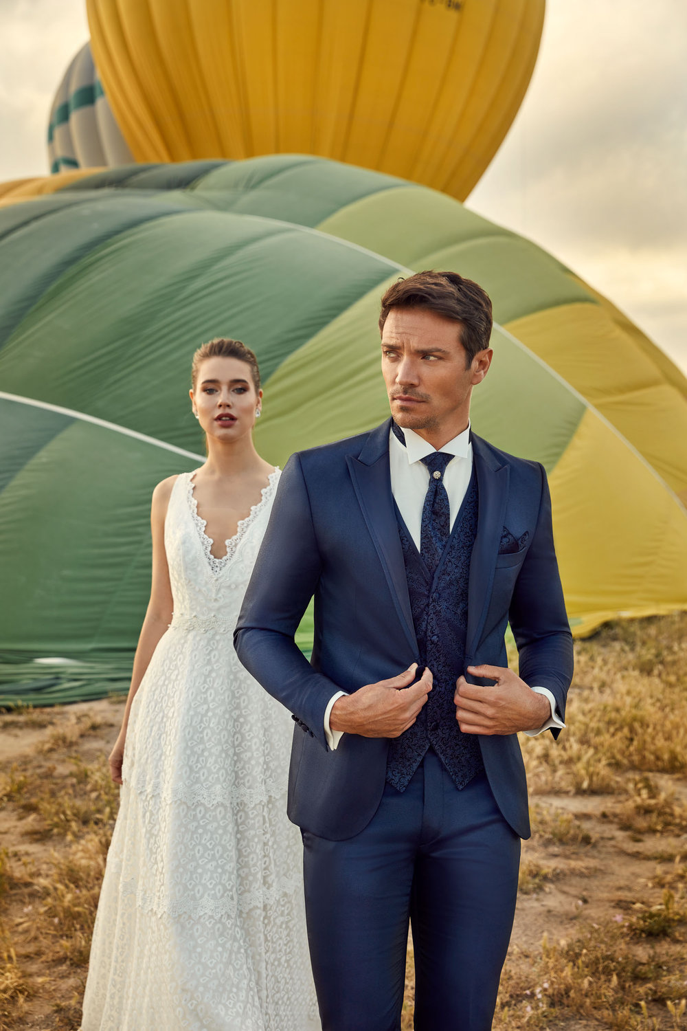 Detailaufnahme eines Bräutigams im blauen Hochzeitsanzug mit taillierter Silhouette und strukturierter Weste. Im Hintergrund ist ein Brautkleid.