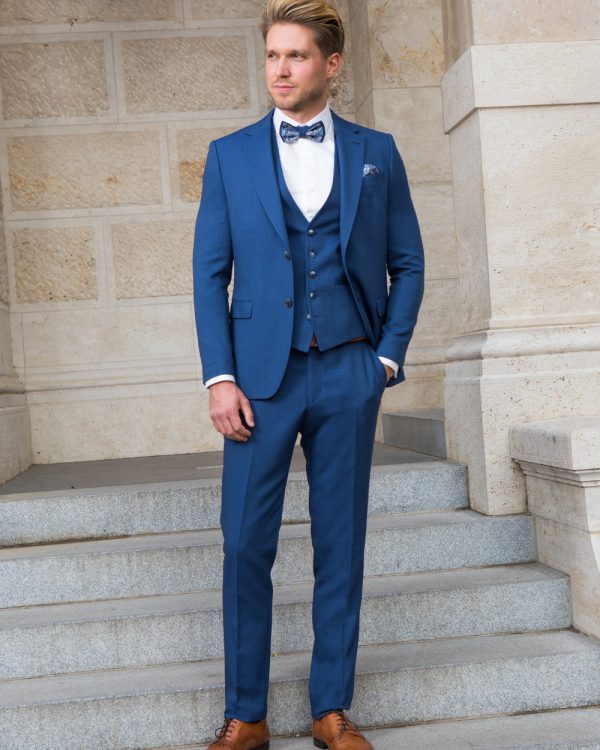 Eleganter dreiteiliger Bräutigamanzug in Königsblau mit Fliege und braunen Lederschuhen, getragen von einem stilvollen Herren auf Steintreppen.
