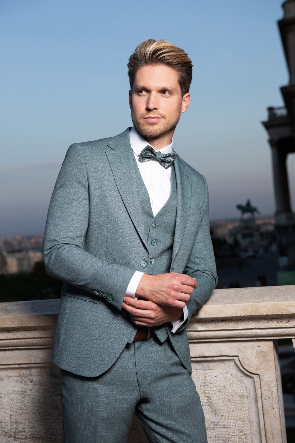 Moderner, hellgrauer Hochzeitsanzug für den Bräutigam mit Fliege und Weste vor romantischem Panorama bei Sonnenuntergang.