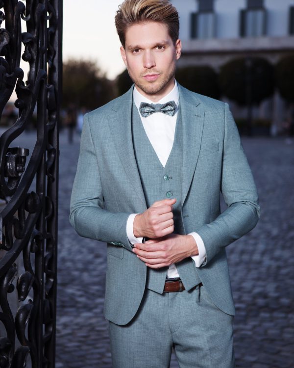 Stilvoller, hellgrauer Bräutigamanzug mit Weste und Fliege, getragen von einem eleganten Herren bei Dämmerlicht am schmiedeeisernen Tor.