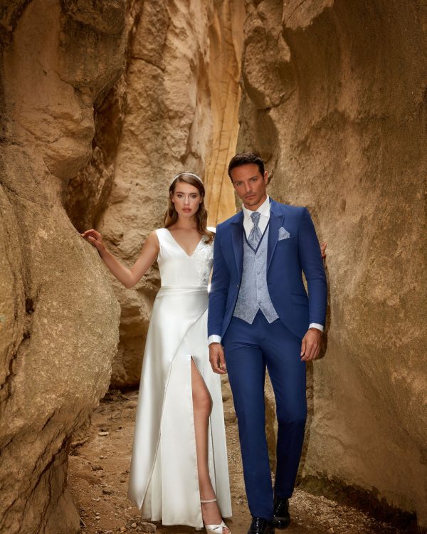 Blauer Bräutigamanzug mit hellgrauer Weste und Krawatte – stilvoller Hochzeitslook in Kombination mit weißem Brautkleid in Felskulisse.