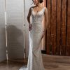 Elegantes Fit & Flare Brautkleid mit tiefem V-Ausschnitt, glitzernder Spitze und langer Schleppe – aufgenommen in stilvoller Indoor-Kulisse mit rustikaler Holzwand.