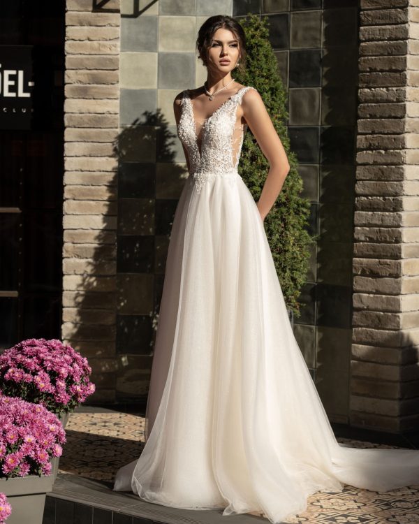 Braut in einem eleganten A-Linie Brautkleid mit tiefem V-Ausschnitt, floraler Tattoo-Spitze und transparentem Tüllrock, stehend vor einer Steinwand im Tageslicht, romantischer Look mit natürlichem Lichtspiel.