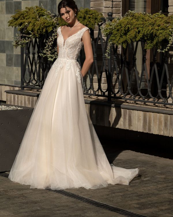 Vollansicht eines eleganten A-Linie Brautkleids mit tiefem V-Ausschnitt, zartem Spitzentop, fließendem Tüllrock und tiefem Rückenausschnitt – fotografiert im Tageslicht vor stilvoller Kulisse.