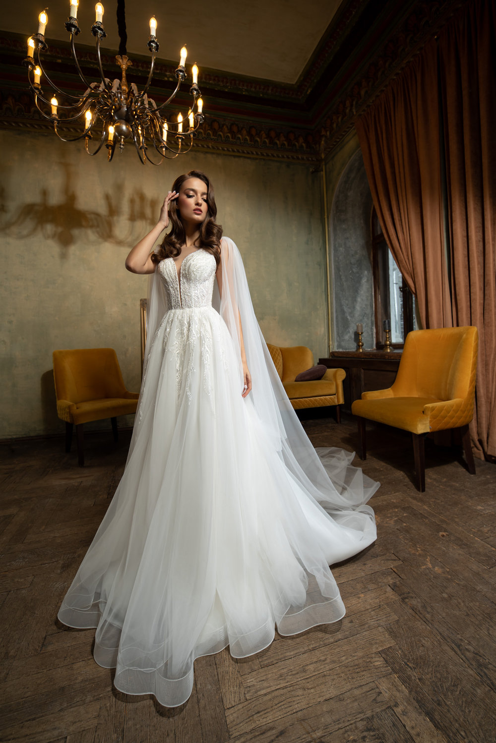 Elegantes Prinzessin Brautkleid mit fließendem Rock, transparenter Corsage und romantischen Cape-Ärmeln – fotografiert in einem stilvollen Vintage-Ambiente mit Kronleuchter.