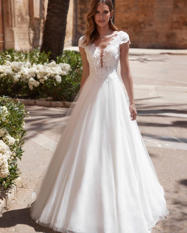 Braut in einem eleganten Brautkleid mit floraler Spitze, V-Ausschnitt und fließendem Tüllrock, stehend in sonnigem Außenbereich vor Blumenbeeten