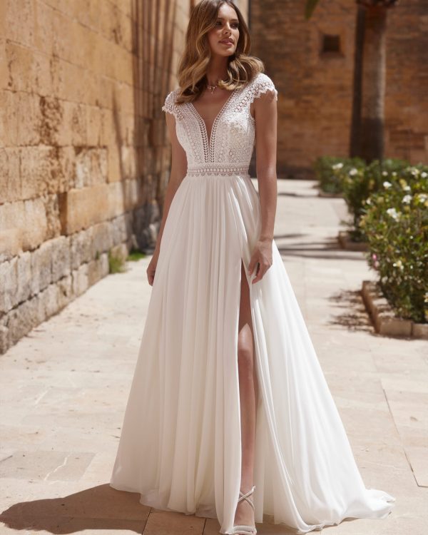 Braut in einem modernen Brautkleid mit tiefem V-Ausschnitt, Cap-Ärmeln und hohem Beinschlitz, stehend vor einer histirischen Steinmauer