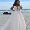 Curvy Brautkleid mit tiefem V-Ausschnitt, floraler Spitze und Tüllrock, getragen am Strand vor dem Meer