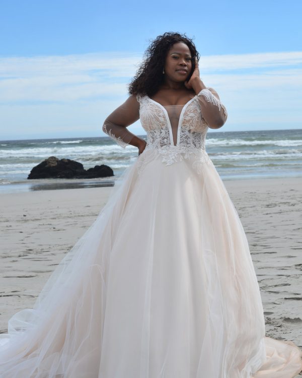 Curvy Brautkleid mit tiefem V-Ausschnitt, floraler Spitze und Tüllrock, getragen am Strand vor dem Meer