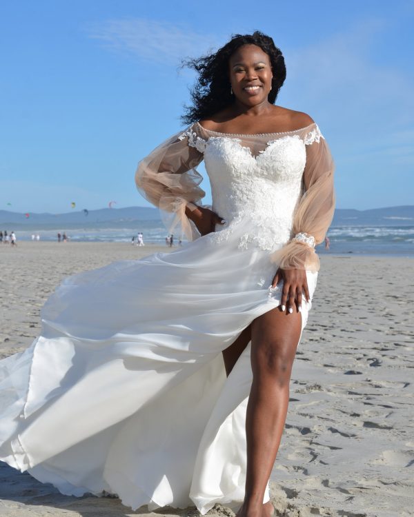 Curvy Brautkleid mit sinnlichem Beinschlitz, langen transparenten Ärmeln und schulterfreiem Spitzenmieder am Strand bei blauem Himmel.