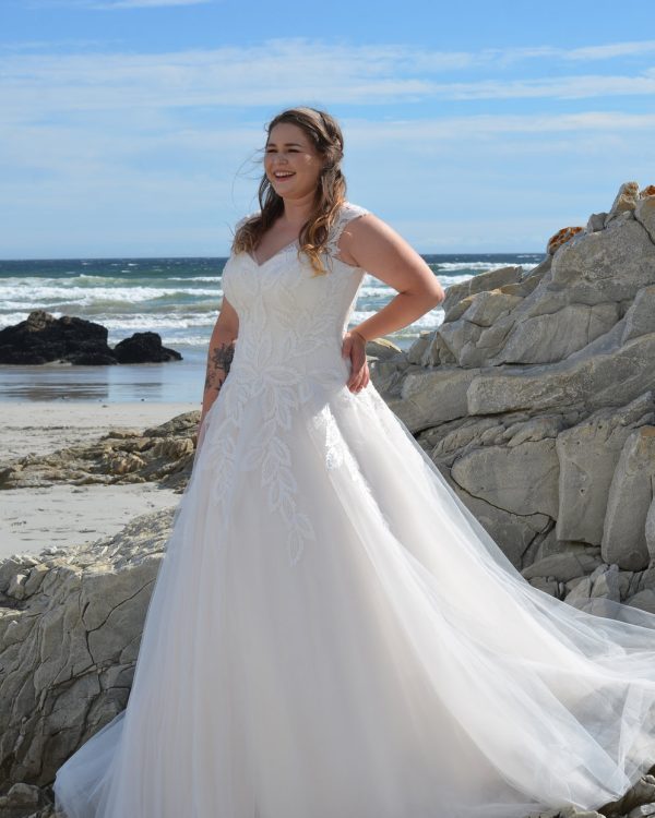 Plus Size Brautkleid mit herzförmigem Ausschnitt, floraler Spitze und Tüllrock, fotografiert am Strand vor Felsen und Meer