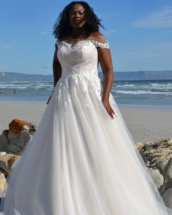 Braut in einem A-Linien-Brautkleid mit Carmen-Ausschnitt und floraler Spitze, stehend am Strand vor dem Meer unter blauem Himmel.