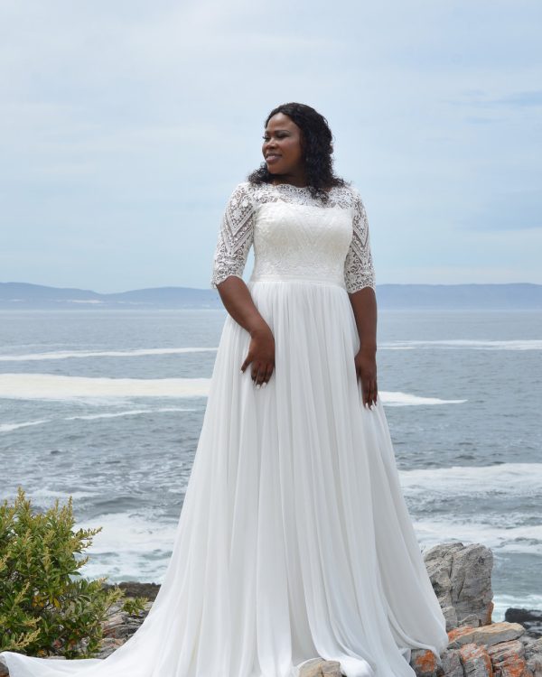 Braut in einem eleganten, langärmligen A-Linien-Brautkleid mit Spitze und fließendem Rock, stehend auf einem Felsen mit Blick auf das Meer.