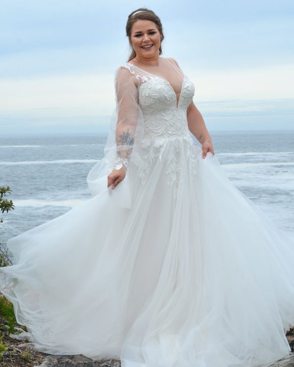 Lachende Braut in einem glitzernden A-Linien-Brautkleid mit tiefem Ausschnitt und Tüllrock, stehend am Meer vor türkisblauem Wasser.