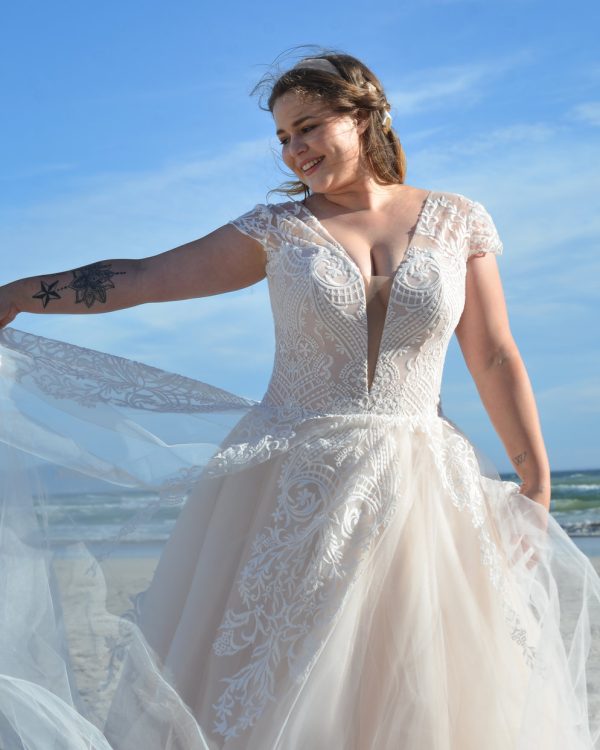 Lächelnde Braut in einem figurbetonten Brautkleid mit tiefem V-Ausschnitt, floraler Spitze und Nude-Schimmer, posierend am Meer unter blauem Himmel.