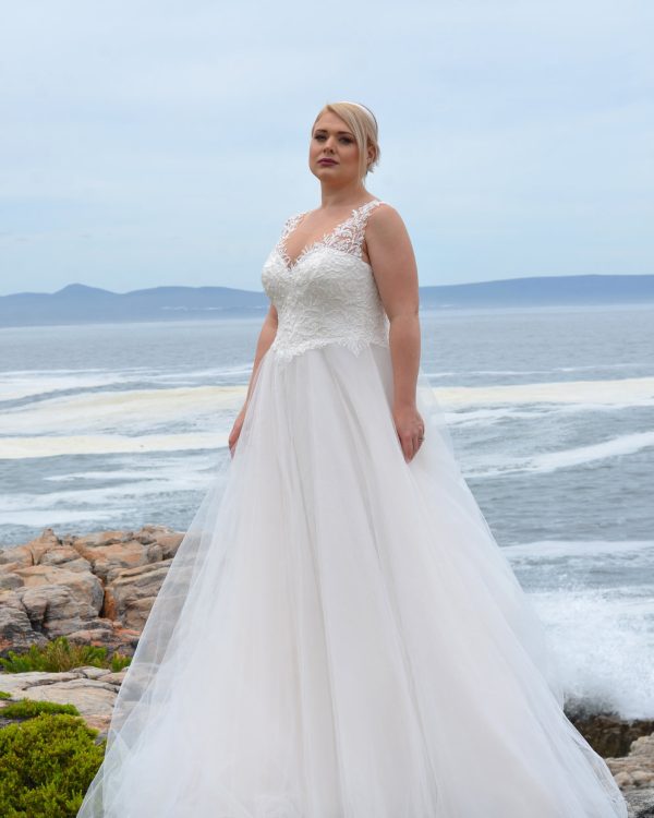 Braut in einem eleganten A-Linien-Brautkleid mit Spitzenträgern und Tüllrock, stehend auf Felsen vor dem Meer bei klarem Himmel.