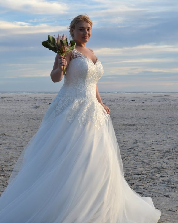 Braut in einem figurbetonten Curvy-Brautkleid mit Spitzenkorsage und fließendem Tüllrock am Strand bei Sonnenuntergang.