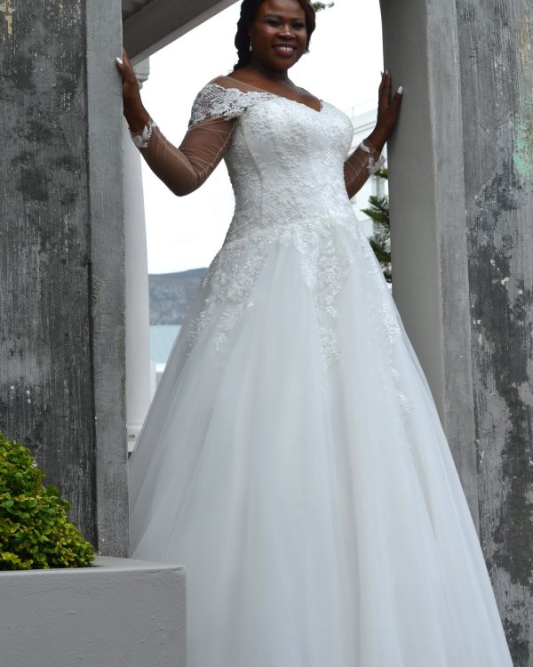 Braut in einem romantischen Curvy-Brautkleid mit Spitzenkorsage, Carmen-Ausschnitt und fließendem Tüllrock, stehend zwischen zwei Steinbögen.