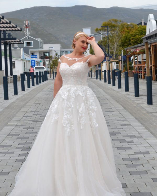 Braut in einem eleganten A-Linien-Brautkleid mit transparentem Illusionsausschnitt und floraler Spitze, posierend auf einer gepflasterten Straße mit Bergkulisse.