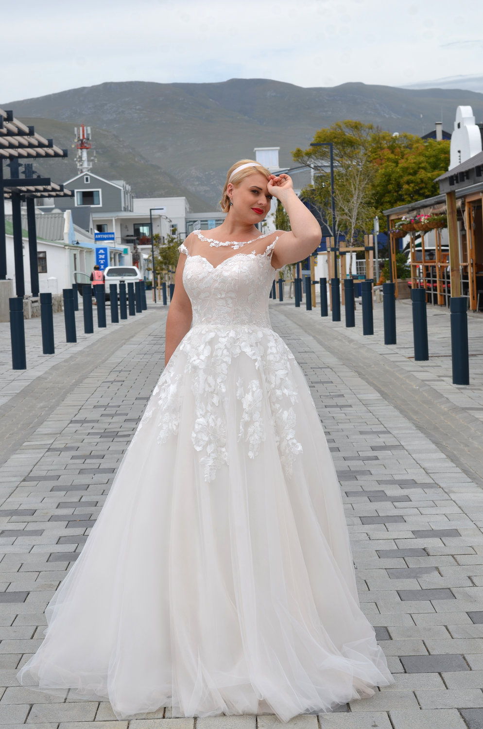 Braut in einem eleganten A-Linien-Brautkleid mit transparentem Illusionsausschnitt und floraler Spitze, posierend auf einer gepflasterten Straße mit Bergkulisse.