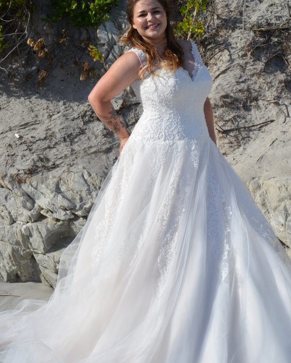 Strahlende Braut in figurbetontem Spitzen-Brautkleid mit V-Ausschnitt am Strand bei Sonnenlicht.