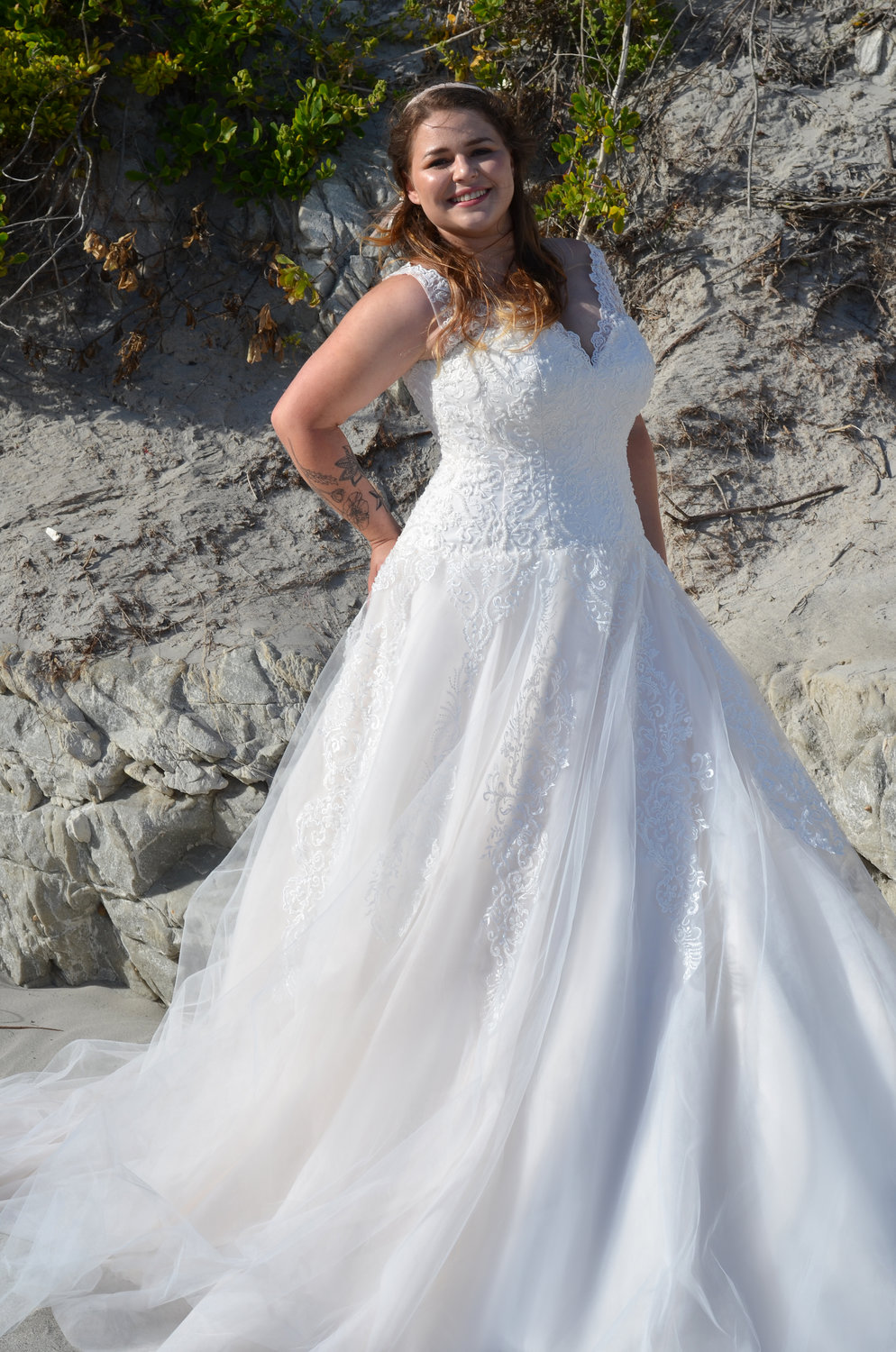 Strahlende Braut in figurbetontem Spitzen-Brautkleid mit V-Ausschnitt am Strand bei Sonnenlicht.