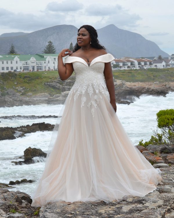 Curvy Braut in einem schulterfreien Brautkleid mit glitzernder A-Linie vor idyllischer Landschaft mit Bergen und Wasser.